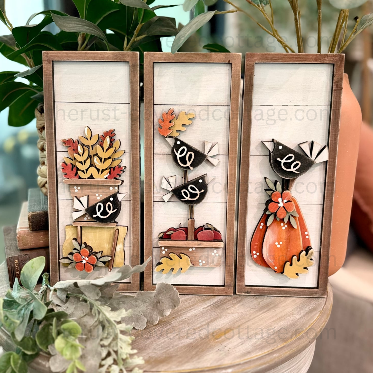 Three vertical wooden decorative panels with layered autumn-themed cutout designs, including birds, pumpkins, leaves, and flowers, displayed on a wooden surface with greenery and a terracotta pot in the background.