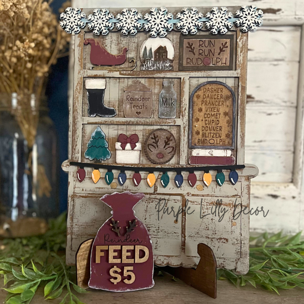 A rustic wooden sign or decorative display featuring a weathered wood panel with small illustrated and text elements, including a red bird, boots, a Christmas tree, and a hanging pennant that reads 'FEED $5'. The item is styled on a tabletop with greenery and a distressed white door in the background.