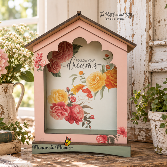 Floral house-shaped sign with “Follow Your Dreams” lettering displayed on worn wood with chippy white architectural accents and soft natural light.