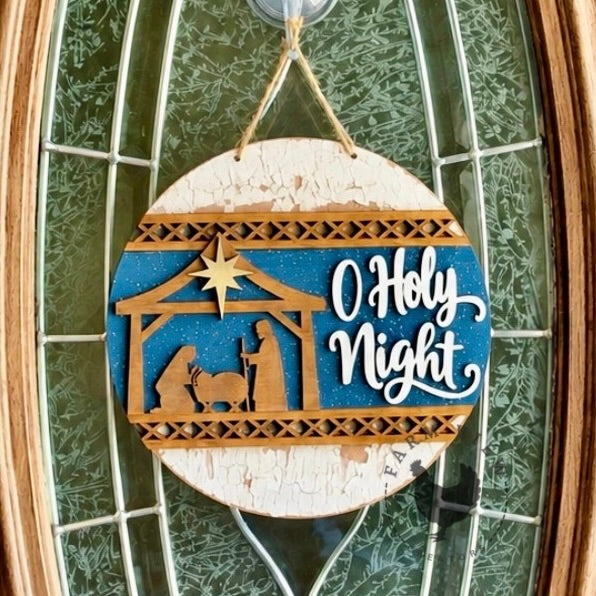 Painted O Holy Night Interchangeable Round Insert with a gold-toned nativity, white lettering, and blue background on a distressed white round, hanging on a glass door.