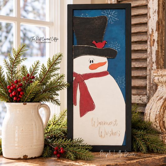 Warmest Wishes snowman sign displayed on a worn wood cabinet with evergreen sprigs, red berries, a crock vase, and salvaged architectural wood in warm natural light.